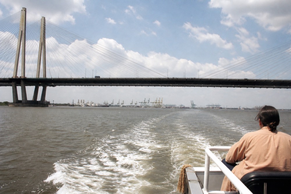 A riverboat passing under a large bridge on the Savannah River with port cranes in the distance, no text, no logos, no people
