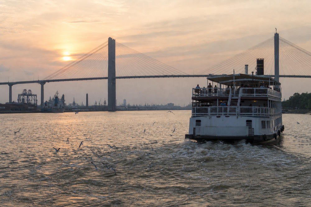 A riverboat on the Savannah River with the sun low behind the bridge cables, no text, no logos, no people