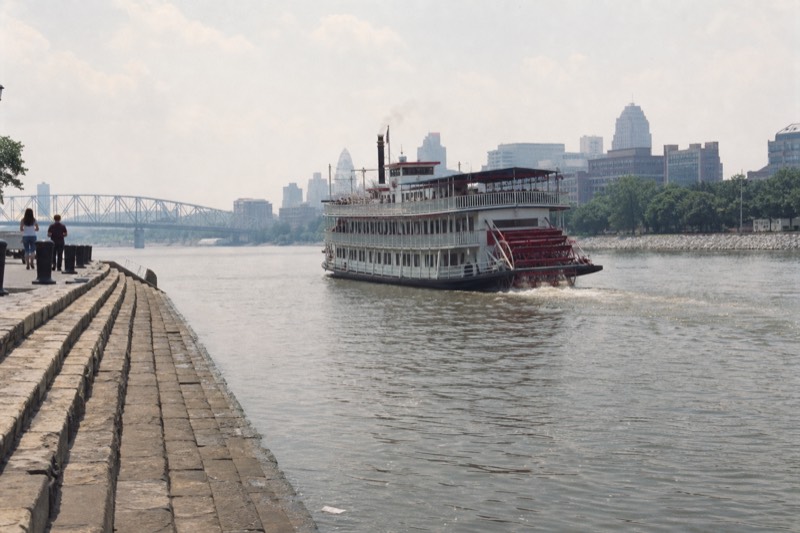 A narrated riverboat easing past Savannah's historic brick riverfront warehouses on River Street under midday light, departing from the historic River Street waterfront in downtown Savannah